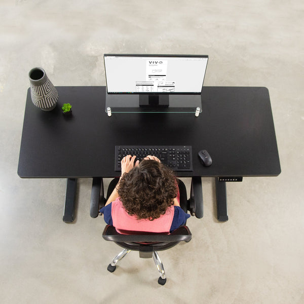 The image shows a birds eye view of a woman typing on the desk, using a computer and looking at a screen.